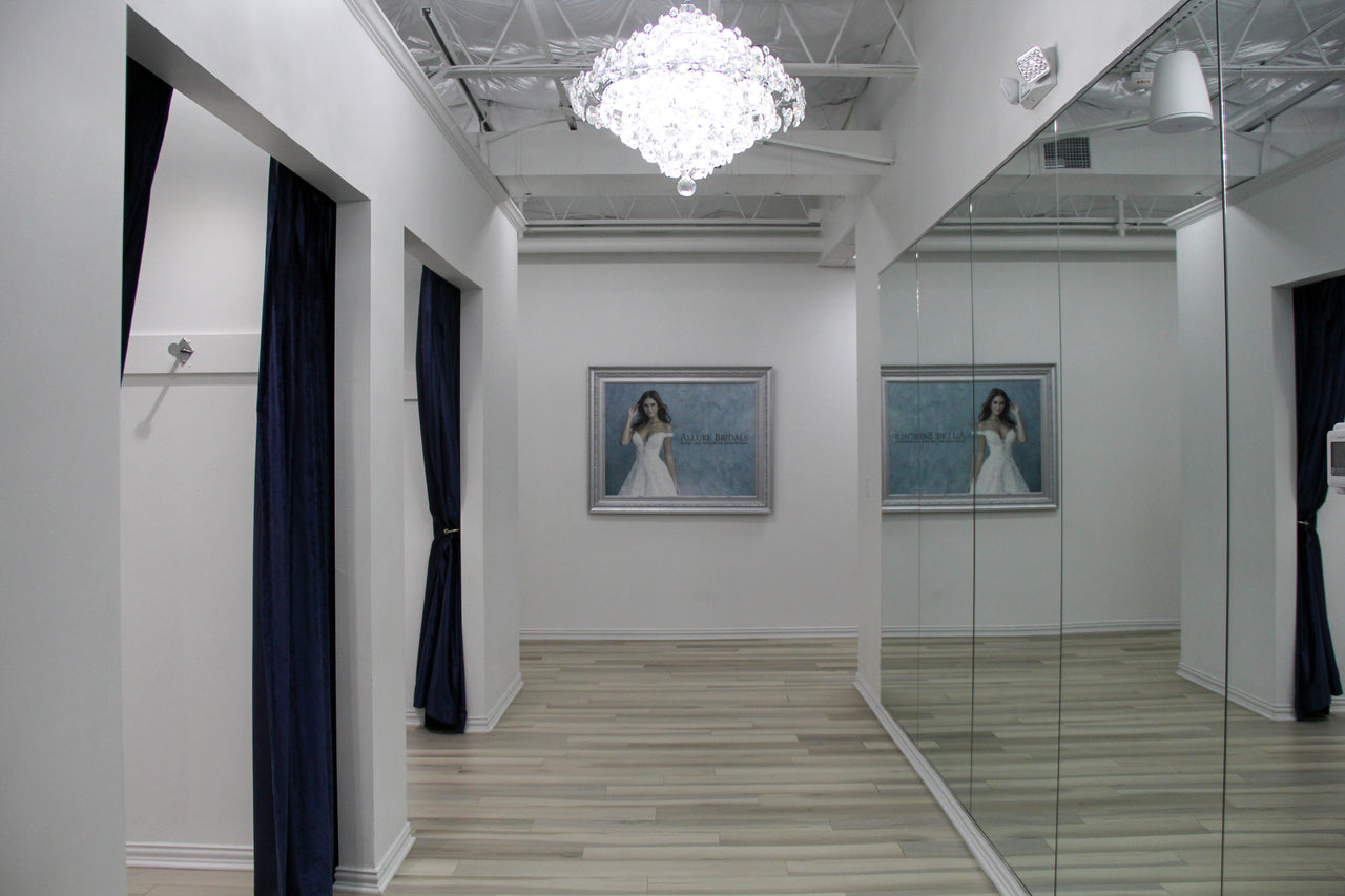 Bridal boutique fitting room area with elegant chandelier, floor-length mirrors, and dressing rooms with navy curtains.