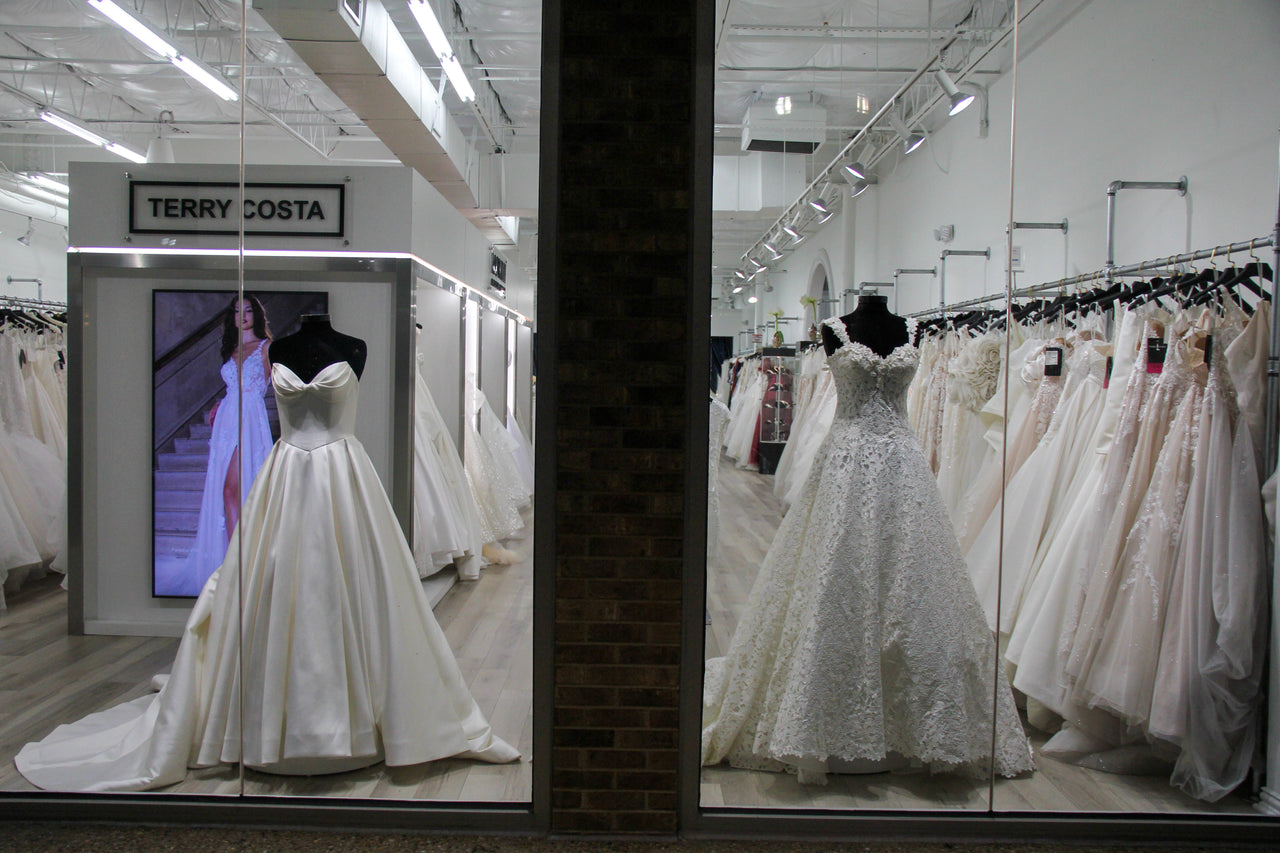 Bridal boutique window display featuring satin ballgown and lace off-the-shoulder wedding dress on mannequins, with racks of gowns inside Terry Costa.