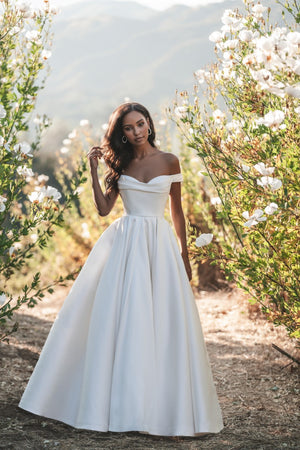 Off-the-shoulder satin A-line wedding dress with pleated bodice and full skirt, styled outdoors in nature.